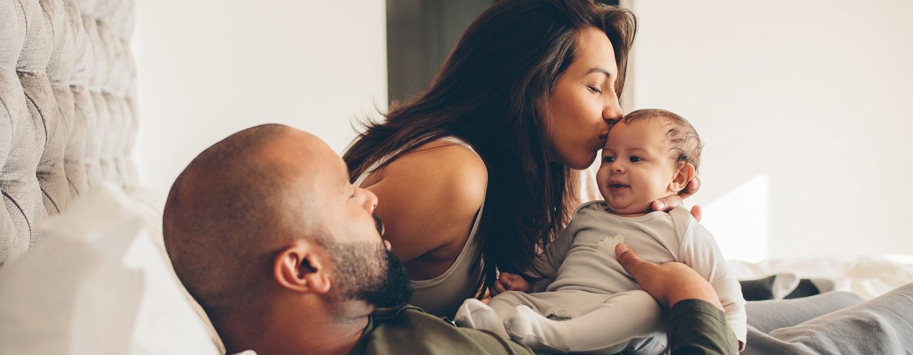 mother kisses her infant while father lies in bed and holds him